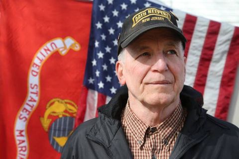 An elderly caucasian man neatly dressed in a dark jacket and Oath Keepers ballcap stands in front of flags for the Marine Corp and USA at an outdoor event
