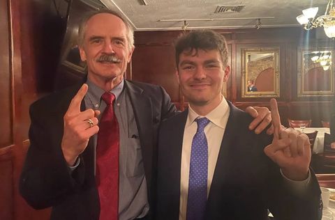 Jared Taylor and Nicholas Fuentes posing for a photo in a room with hardwood walls and a chandelier, both men are in a suit and tie and holding up their index finger pointing up.