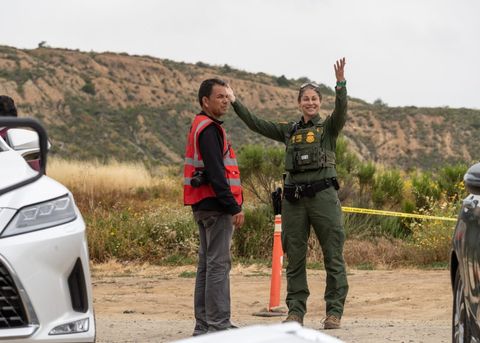 a border patrol agent looks at the camera and throws her hands in the air, smiling while a man in a vest with a camera stands next to her looking away, his hands at his sides