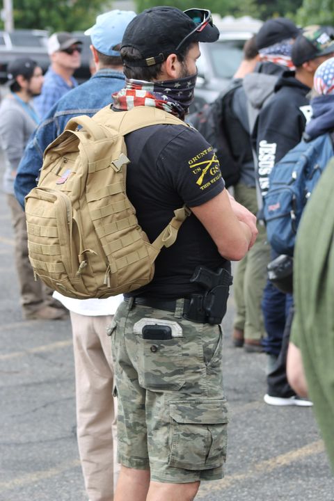 A Proud Boy in camo shorts with a baseball cap and a handgun holstered on his hip.