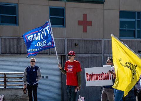 a red cross is seen on a wall in the background, in the foreground a mask-less trump supporter wearing a red trump 2020 shirt, and red hat holds up a trump 2020 flag with one hand, and is wearing dark sunglasses to the right of that person is a white sign with red text that reads 'Brainwashed!' and the the right of that sign is a don't tread on me flag