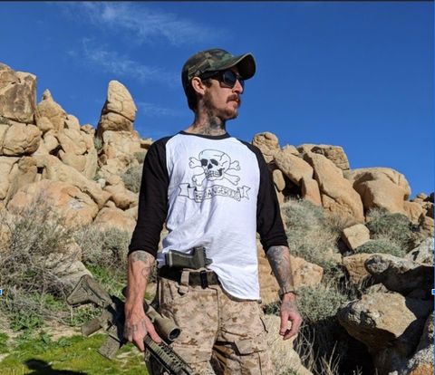 A man poses in the desert with a camoflauge-painted rifle in one hand and a gun tucked into his waistband. He's wearing a shirt with a totenkopf, a stylized a skull and crossbones nazi soldiers would wear.