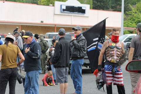 Three men in body armor, sunglasses and black jackets, one holding a flag in a crowd of similar men. Off to the side is a man wearing a red gaiter over the bottom half of his face with fake Roman soldier armor with the words 'cucked' written over it with stickers