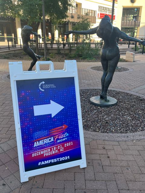 An A frame sign with an arrow points in the direction of TPUSA’s AmericaFest. Sign reads 'Turning Point USA. America Fest 2021, A Turning Point USA Event. December 18 to 21 2021 Phoenix, AZ. hashtag AMFEST2021' The sign is placed in from of two nude statues in the nearby area