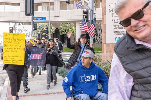 An old man in sunglasses grimaces in front of the camera to the right as a crowd with a banner that says "lexit" waves signs and american flags walking down the street in front of a childrens' hospital