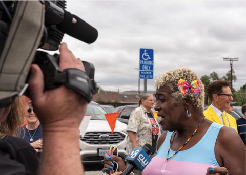 A woman with earrings and a rainbow necklace talks to a reporter as a cameraman films her