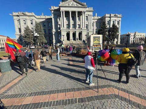 Counter protestors stand in front of the capitol before Westboro Baptist arrives rainbow and red flags are seen. People are hanging out preparing for WBC to arrive. Two people are doing an interview holding up rainbow umbrellas