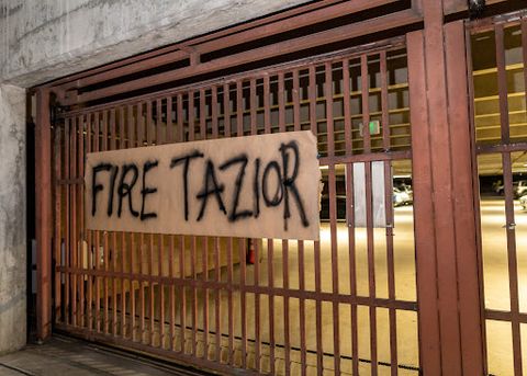 a paper sign is taped on the police department’s parking garage gate. The sign is made of brown paper, and the message 'Fire Tazior' has been spray painted on it in black.
