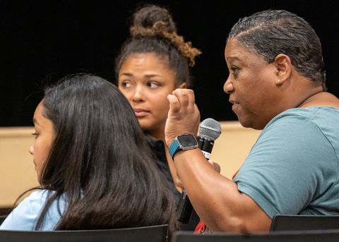 Tasha Williamson is on the right side of the frame, shot from the ribs up in profile, sitting. She is gesticulating and speaking into a microphone, looking at the panelists with a stern expression.