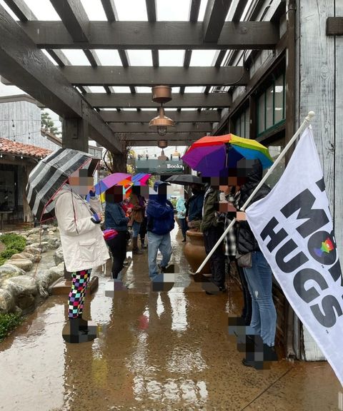 a bunch of parents standing around a bookstore in rain gear with rainbow umbrellas. one holds a sign that says "free mom hugs." Their faces and shoes are blurred out to protect their privacy