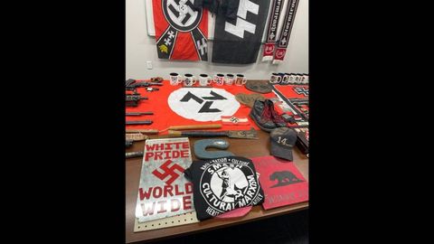 A collection of Nazi memorobilia on a table including a shirt that says "smash cultural marxism," a triskele nazi flag, a nazi armband, nazi coffee mugs and boots with red laces