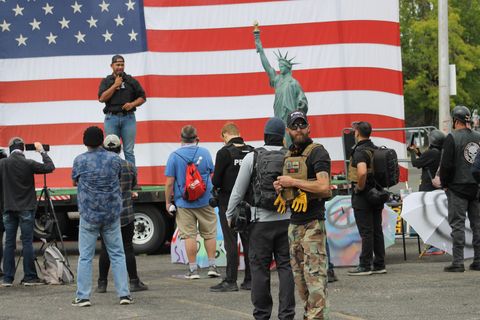 A tall man stands with a microphone on a stage with a giant american flag behind him with a person-sized statue of liberty model off to the side while men in body armor mill about