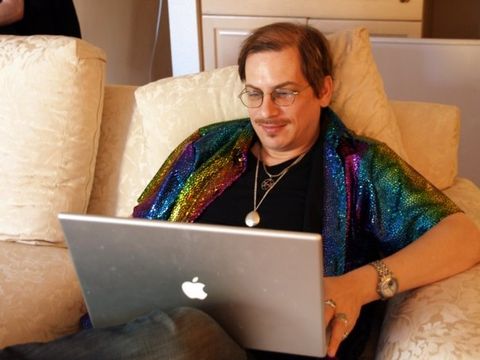 Horat is visible from the waist up, sitting on a couch with one leg crossed to rest its foot on the opposite knee. He is using a Macbook, he has numerous rings on the visible hand, and a rainbow-sequin shirt. He is looking down at his laptop, smirking slightly. He is wearing glasses, has a soul patch, and a mustache. He has two visible necklaces, one of which is Crowley's unicursal hexagram.