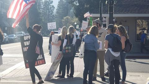 people standing around on a street corner waving american flags while some chat, too tired to hold up their signs anymore