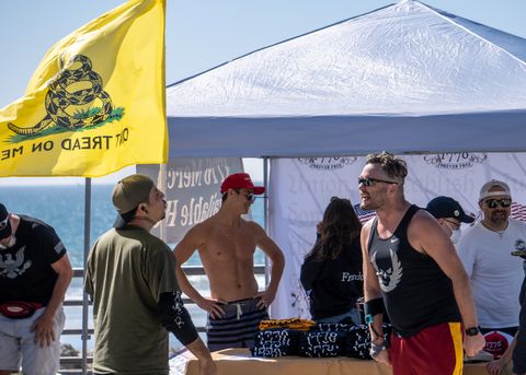 In the foreground there are two men who appear to be shouting aggressively at eachother, neither has a mask on, and the one on the left is wearing a matching olive green hat and t shirt with a long sleeve black shirt underneath. he has a band neck tattoo. the other man is wearing a black tanktop and running shorts, with sunglasses on. behind them there is a dont tread on me flag, and a pop up tent with what appears to be merch, and other maskless people milling about.