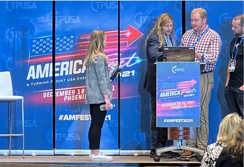 Christian musician and failed congressional candidate Sean Feucht prepares to deliver a speech. Three people stand on stage helping him set up his computer. The backdrop behind Feucht and the sign in front of him both advertise AmericaFest.