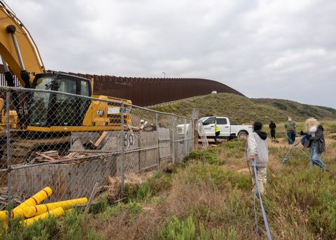 On the left side of the frame, an excavator sits behind a fence. On the right hand side, protesters walk away from the camera holding a rope. Beyond them, Spencer Construction employees are getting into their truck. The border wall is visible in the background.