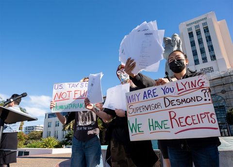 Protesters are large in the frame, with blue skies and the county administration building behind them. Protesters have fanned out receipts to show as many as possible in one hand. The three protesters have two signs among them. One reads, "why are you lying supervisor desmond?! we have receipts." The other reads, "US Tax dollars do not fund humanitarian aid," but it's partially obscured by another protester.