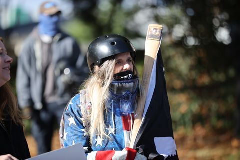 Cindy Loren, who based on her attire, believe she's some sort of MAGA super solider, can be seen in this photo. She's wearing a black skateboard helmet, a low-slung mask not even covering her nose, and a respirator off her neck. She's wrapped in an American Flag and wields what appears to be an axe handle but without the blade.