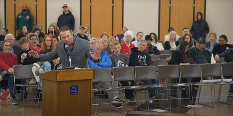 This is a screenshot of the live feed from the Oceanside Unified School District Board of Education meeting on February 7th, 2023. In it, Bryce Henson can be seen commenting to the board, all of whom are off-screen. Henson is standing at a podium and gesticulating. There are numerous attendees out of focus behind him.