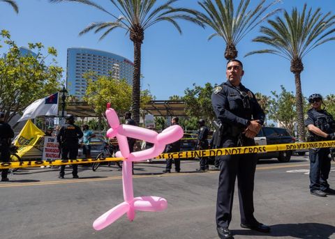 Police formed perimeters on either side of the street outside the New Children's Museum on August 5th. Someone tied a balloon animal to the caution tape. A pink balloon animal is in the foreground, and caution tape runs across the middle of the frame. Behind the tape, two officers stand facing the viewer. Across the street, officers stand with their backs facing the viewer. The officers are facing a crowd of protesters, one of whom has a Christian flag, and another of who has a Don't Tread on Me flag.