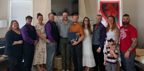10 white adults and one child (censored) posing for a photo at the first Raven Folk United meetup. A red and black raven flag is pinned behind them. A flatscreen also sits on the wall behind them. Krogstad is wearing a white dress and standing next to Drago who is wearing a long-sleeved purple button up shirt, an ugly patterned vest, red tie and Thor’s Hammer necklace. Other men in the group are wearing a similar getup.