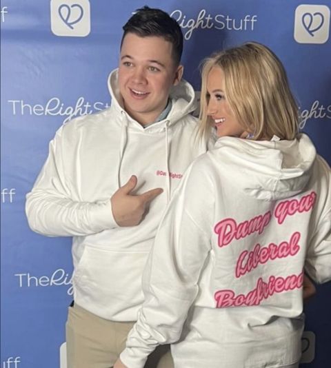 Kyle Rittenhouse and his blonde girlfriend Skyler Bergoon pose in front of a light blue ad wall for The Right Stuff dating app. Both are wearing white pullover sweaters that read “Dump you liberal boyfriend” in pink cursive text. The front reads “# Date Right Stuff” Both are smiling. Kyle points to the text on his sweater.