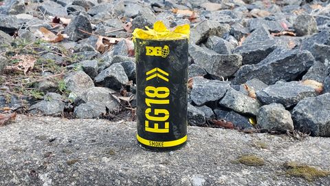 A black canister with yellow text standing upright on the curb. Behind it are a bunch of jagged grey rocks. The canister has yellow text and says EG18 and SMOKE. At the top is the Enola Gaye logo. There is the paper insert and powder visible on top as it is open.