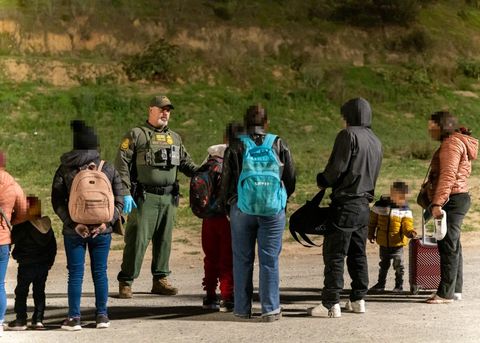 Parents and children stand, facing a border patrol agent. Some of the children are waist-height, and all asylum seeker are wearing multiple layers to withstand the cold. The background consists of splotchy grass and dirt. It is night, and the subjects are lit by artificial light.