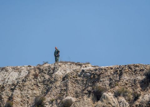 A Border Patrol agent stands on a stony, tan ridge. The sky is blue. He is small in the frame, visibly distant from the camera.