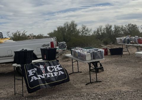 a Truck parked in the dirt along the side of the road has set up a flag merchandise booth, one flag on display reads 'Fuck Antifa' the 'U' in 'Fuck' is a masked skull that has 'Est. 1775' stamped on it’s forehead.