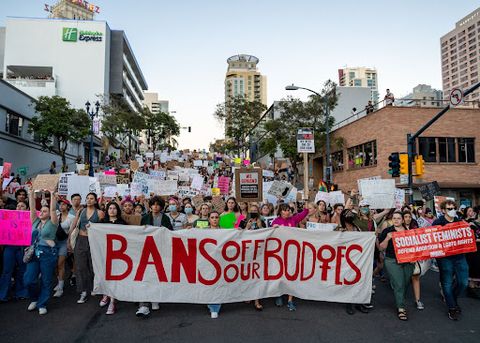 Thousands of protesters march through downtown San Diego at dusk, occupying the breadth of the street. A banner is held by six people at the front of the crowd which reads, “Bans Off Our Bodies.” 