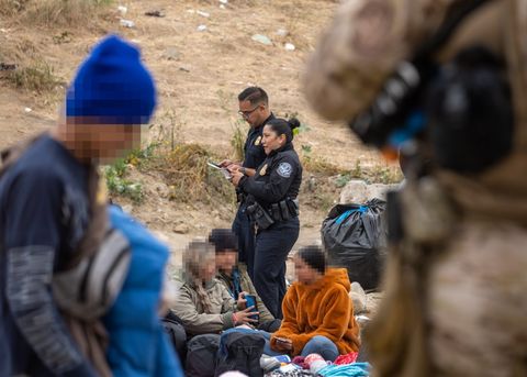 two border patrol agents look at clipboards as they stand by people sitting on the ground