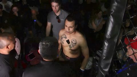 A shirtless man with a piece of tape over part of his chest covering a tattoo stands in the middle of the crowd before a fight.