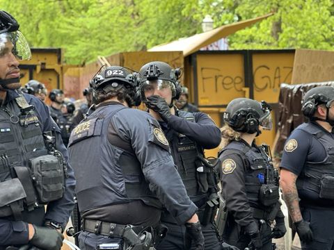 a police officer covers their mouth while whispering as another leans their ear in to listen and others stand around in riot gear