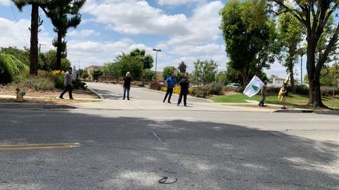 Men in masks standing around the vehicle entrance to a park looking around. one holds a white flag with a green pine tree on it that says 'an appeal to heaven' on it