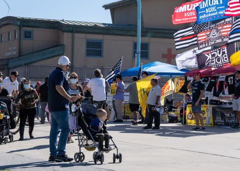 A toddler looks on at the display, while two men with bullhorns take turns comparing the governor to various dictators throughout history. Among the flags for sale is a blue Trump 2024 flag, with the text 'Take America Back' just below the intended year. At the time of writing, Trump has not announced his candidacy.