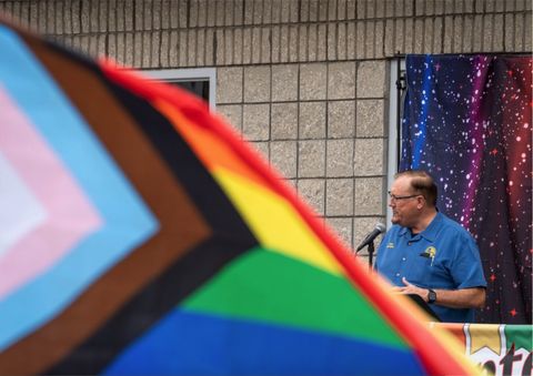 A middle-aged man with glasses stands at a podium in front of a waving pride flag and speaks into a microphone