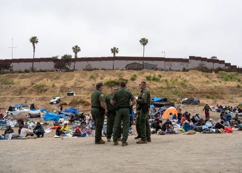 a group of border patrol agents standing around talking next to people sitting on the ground