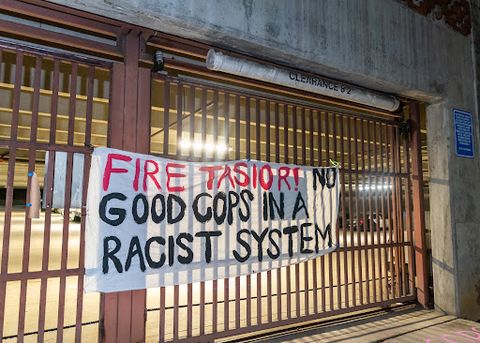 A white cloth banner hangs from the police department’s parking garage gate. In red text, it says 'Fire Tasior!' In black text, it says 'No Good Cops In A Racist System.' The picture is taken at night, but the yellow light of the garage colors the photo