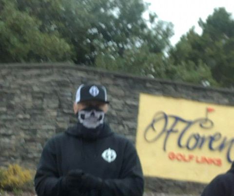 Neo-Nazis look towards the camera. The person at left is largely cut off. Directly in the middle is a man with his face covered holding a white flag. At right is a man, face unobstructed, wearing a white hat. He's flexing hard and locking his jaw as he glares into the camera.
