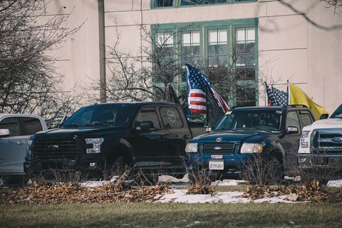 A row of big trucks with various flags on the back and one without a front license plate