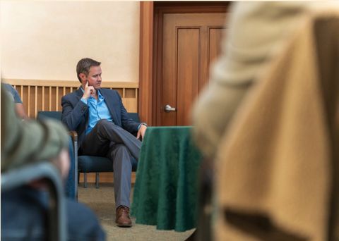 Seth Hall attended the May15h TRUST SD summit and offered commentary. He is sitting, wearing grey slacks and a navy-blue blazer. He is leaning slightly, placing a hand to his jaw as he listens to someone out of frame.
