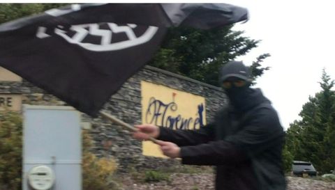 A neo-Nazi in black clothing with his face obscured waves flag with the black sun towards the camera. Behind him is the sign for the Florence Golf Links
