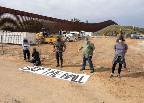 Five protesters stand between the camera and the resource site. A banner that reads "STOP THE WALL" is on the ground in front of them. Numerous construction vehicles and heavy equipment are visible in the background. The border wall extends over the horizon.