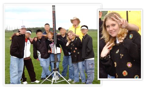 A group of nine people are posing for a picture in an open field. Directly in front of them in the center of the image is a tall, thin rocket. Dallas Humber is the second from the right. She’s wearing blue jeans and a black sweater adorned with gold appliques and oversized multi-colored jewels. She has shoulder length blonde hair and is making a double handed “victory” gesture. 