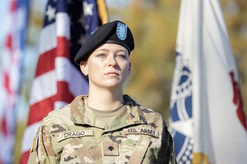 Ashley Krogstad Drago wearing her camouflage uniform and black beret hat. She is standing in front of the American flag. Her tags read “DRAGO” and “U.S. ARMY.”

