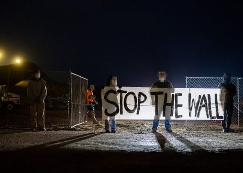 Four protesters stand in frame, holding up a banner that reads "STOP THE WALL." They are backlit by car headlights before dawn. Behind them, a private security employee closes the fence.