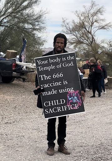 A man holding up a sign that reads 'Your body is the Temple of God. The 666 vaccine is made using CHILD SACRIFICE.' An image of Baphomet is stamped on the bottom.