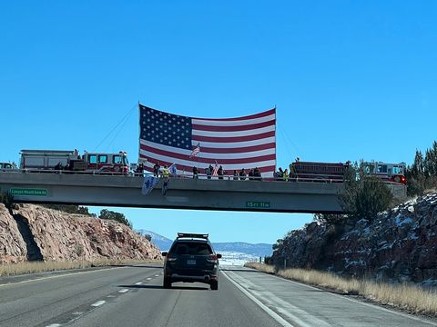 two fire engines straddle a bridge with a giant american flag between them while others on the bridge wave smaller flags and cars on the road pass under